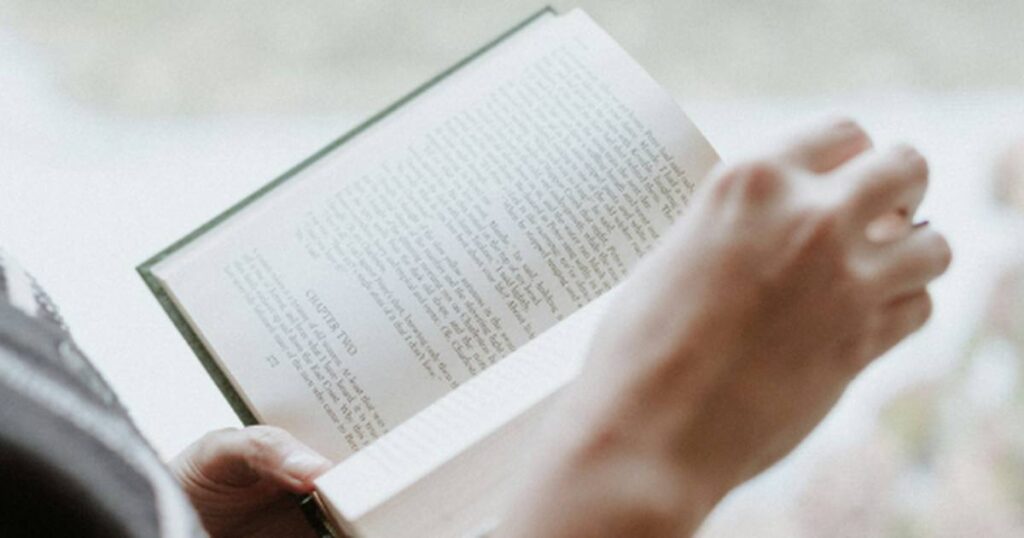 Persona leyendo un libro junto a una ventana con luz natural, símbolo de reflexión y profundización en la fe.